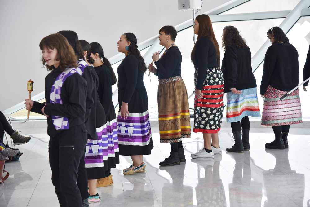 Nine females in ribbon shirts and skirts, singing as they perform an Earth Dance at the Milwaukee Art Museum.